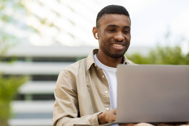İnternete otur. Parkta kulaklıkla otururken dizüstü bilgisayarda yazan gülümseyen siyah adam. Yüksek kalite fotoğraf - Fotoğraf, Görsel