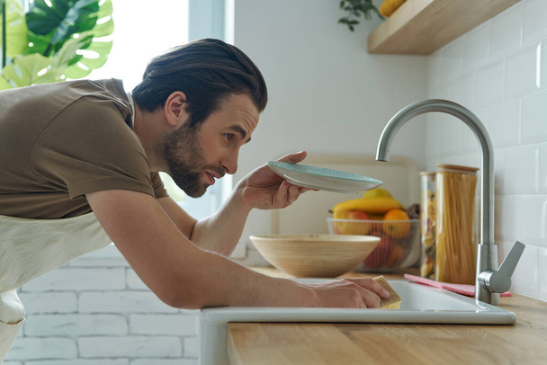 Handsome young man checking dishes after washing while standing near kitchen sink - 写真・画像