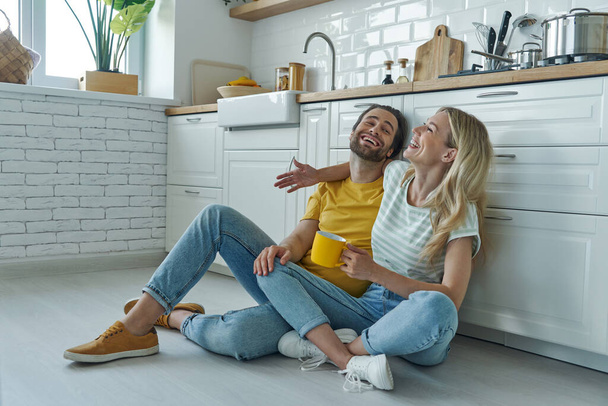 Веселая молодая пара обнимается, сидя на полу и опираясь на кухонный стол - Фото, изображение