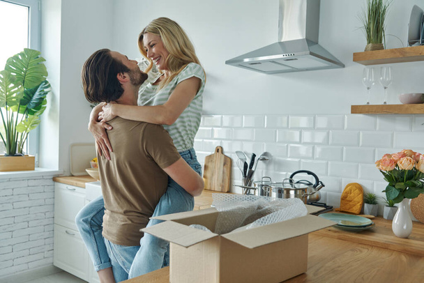 Young couple embracing and smiling while open box laying on the kitchen counter - Фото, изображение
