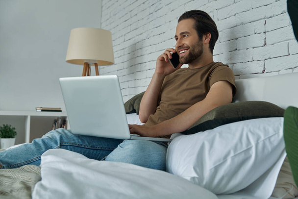 Cheerful young man using laptop and talking on mobile phone while relaxing on bed at home - Φωτογραφία, εικόνα