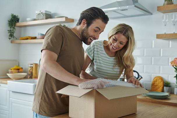 Young couple unpacking box and smiling while standing at the domestic kitchen - Foto, immagini