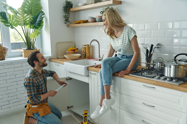 Χαρούμενος νεαρός άνδρας επισκευάζει ένα νεροχύτη ενώ η γυναίκα κάθεται στον πάγκο της κουζίνας - Φωτογραφία, εικόνα