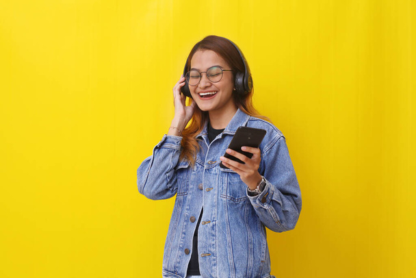 Happy young asian woman standing while listening to music from her phone. Isolated on yellow - Photo, Image