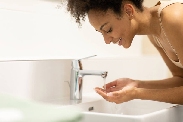 Mujer afroamericana se lava la cara con agua en el lavabo del baño.. Foto de alta calidad - Foto, imagen
