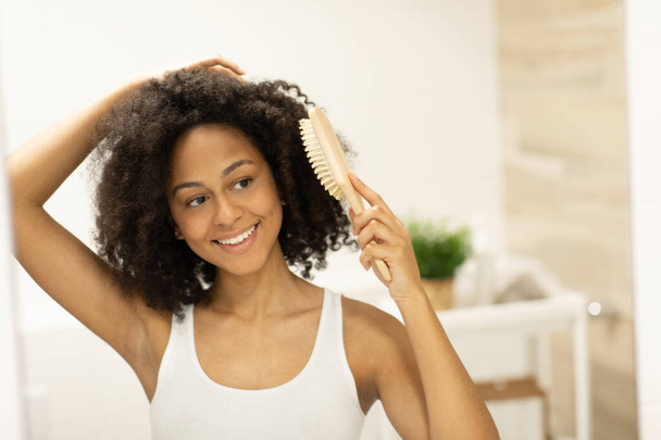 Uma jovem mulher em uma parte superior do tanque fica na frente de um espelho do banheiro e penteia seu cabelo com uma escova de madeira depois de um banho. Foto de alta qualidade - Foto, Imagem