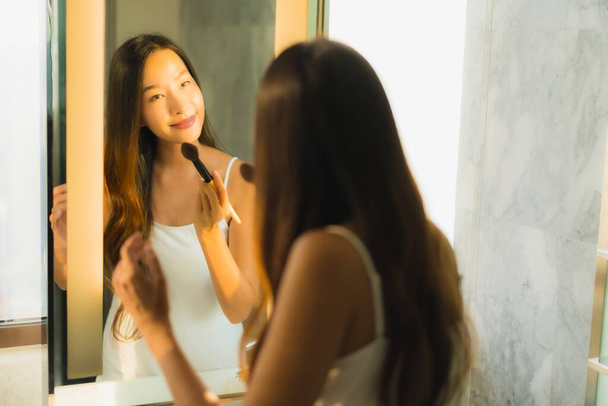 Retrato hermosa joven mujer asiática maquillaje con polvo cosmético y lápiz labial en su cara facial en el interior del baño
 - Foto, imagen