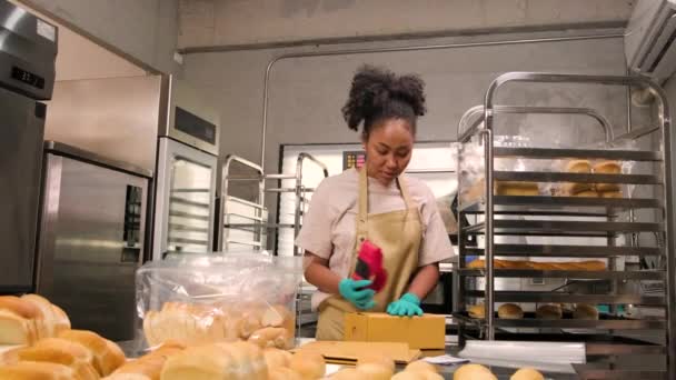 Avvio panetteria consegna piccola impresa. Una cuoca afroamericana sta confezionando pane e dolci fatti a mano e appena sfornati in scatole e inviando per gli acquisti online dei clienti in cucina culinaria. - Filmati, video
