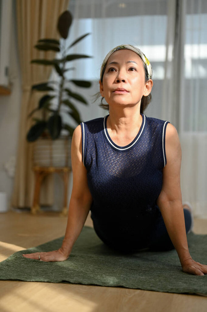 Спортивная и здоровая азиатка в возрасте делает легкие пилатесы дома, растягивая свое тело на коврике для йоги. Пожилые люди с концепцией здравоохранения - Фото, изображение
