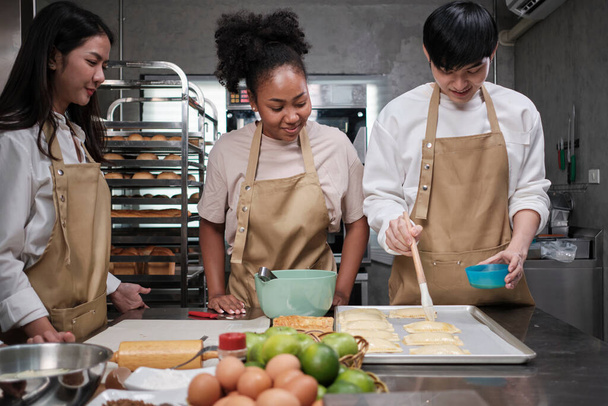 Cuisine course, a young male chef in apron and group of cooking class students, brushes pastry dough with eggs cream, prepares ingredients for bakery foods, fruit pies in stainless steel kitchen. - Foto, Bild