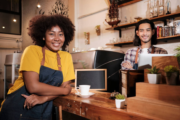 Two cafe business startup partners and friends, African American female, and Thai male baristas look at camera, arms crossed, and cheerful smiles at coffee shop counter bar, happy service staff job. - Photo, Image