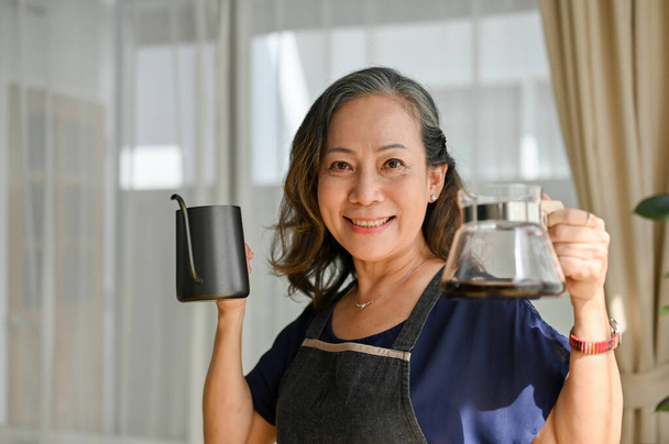 Счастливая и жизнерадостная азиатка в возрасте держит в гостиной чайник с горячей водой и горячим кофе. Пивоварение кофе - Фото, изображение