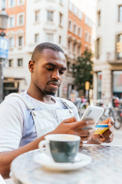 Selbstbewusste junge bärtige Afroamerikaner in lässiger Kleidung. Er sitzt mit einer Tasse Kaffee am Tisch und bedient das Smartphone, während er im Café im Freien mit Kreditkarte bezahlt. - Foto, Bild