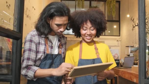 Dva mladí startup barista partneři se zástěrami stojí u příležitostných dveří kavárny, psát na palubě a ukázat otevřenou ceduli, šťastný a veselý úsměv s kavárnou servisní práce, a nové obchodní podnikatelé. - Záběry, video