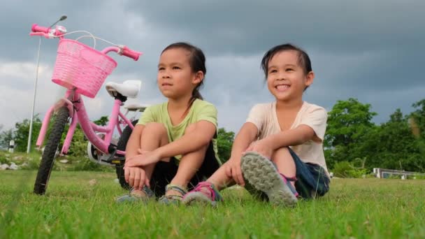 Mutlu küçük kız ve kız kardeşi parktaki bisikletlerin yanında çimlerin üzerinde oturuyorlar. Çocuklar bisikletten sonra dinleniyor. Çocuklar için Sağlıklı Yaz Faaliyetleri - Video, Çekim