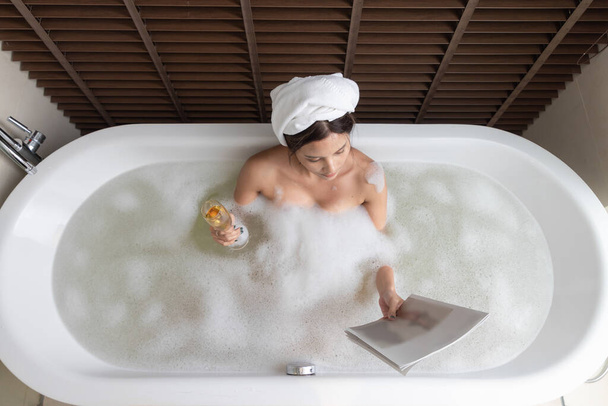 Mulher segurando vidro com vinho branco e deitado cheio de espuma. Relaxar na casa de banho. vista superior - Foto, Imagem