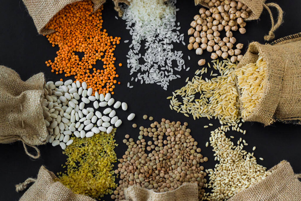 Red lentils, chickpeas, white rice and bulgur pouring from jute sack onto black surface with copy space.Harvest concept - Foto, Imagem