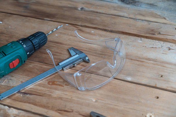 Ηλεκτρικό τρυπάνι, διαμέτρημα, πένσα και προστατευτικά γυαλιά βρίσκονται σε ξύλινη επιφάνεια. Το τρυπάνι είναι τοποθετημένο σε μια τσοκ. Επιλεκτική εστίαση. - Φωτογραφία, εικόνα