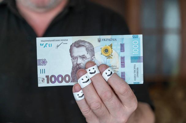 Un uomo adulto tiene i soldi in mano. Mille grivne ucraine. Le prime falangi delle dita sono avvolte in un nastro bianco. Volti sorridenti sono dipinti sulle bende. - Foto, immagini