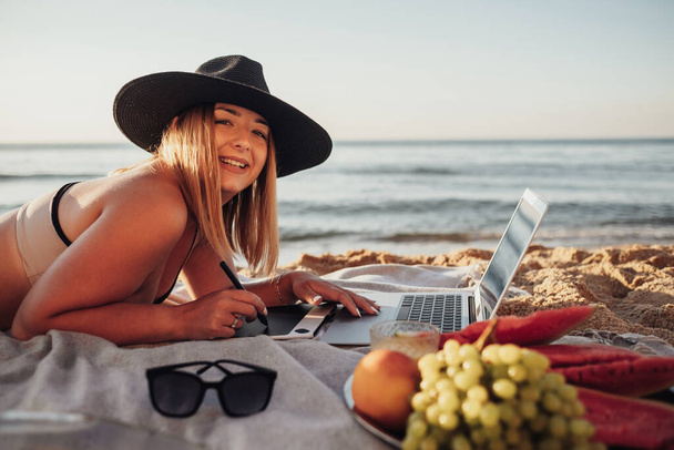 Vista laterale da vicino della donna allegra vestita in costume da bagno che lavora su tablet digitale e laptop durante il picnic via mare - Foto, immagini