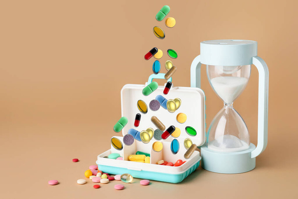 Contenedor con diferentes píldoras y reloj de arena sobre fondo de color - Foto, imagen