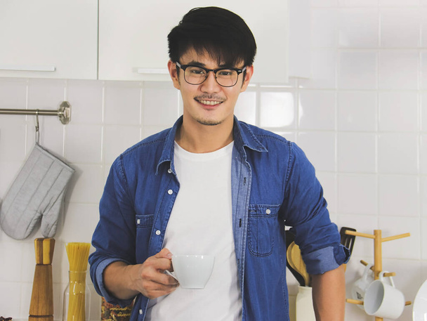 Portrait of young and handsome Asian man wearing eye glasses holding a cup of coffee in the kitchen, smiling and looking at camera. - 写真・画像