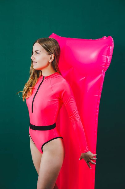 A slender happy woman in a bright pink swimsuit holds and poses with a pink inflatable mattress on a green isolated background. Pink air mattress. Summer pool mattress - Foto, imagen