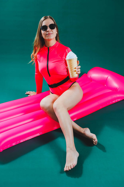Attractive beautiful woman slim body in a bright pink swimsuit in sunglasses lies on an inflatable pink mattress isolated on a green studio background. Summer vacation at the sea, tanning concept. - Photo, Image