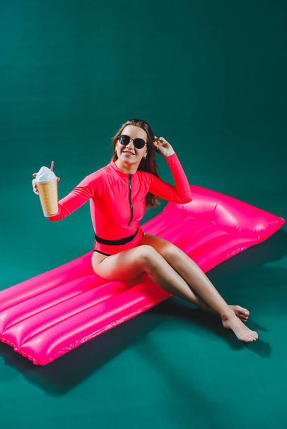 Досить струнка жінка в рожевому купальнику лежить на надувному матраці з коктейлем в руках, на ізольованому зеленому тлі. Літні канікули в концепції морського засмаги. Рожевий повітряний матрац
 - Фото, зображення