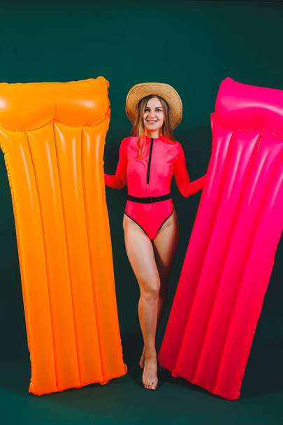 Happy pretty young woman in hat wearing bright pink swimsuit standing over isolated background holding inflatable pink mattress. Inflatable orange mattress. Summer vacation. - Fotografie, Obrázek