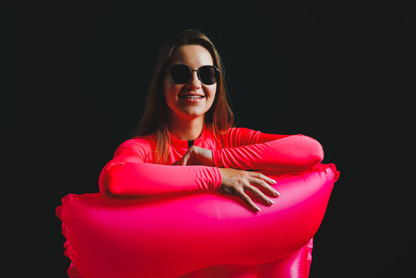 Young blonde woman in a pink swimsuit, isolated on background. Vacation lifestyle concept, summer vacation. A girl in sunglasses holds an inflatable pink mattress in her hands for swimming - Photo, Image