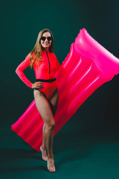 Beautiful woman dressed in pink swimsuit and sunglasses posing with pink inflatable mattress isolated on green background - Foto, Imagem