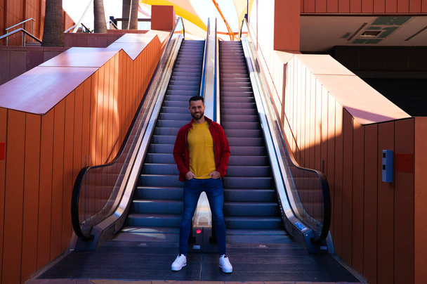 Un beau jeune homme aux yeux bleus et à la barbe se tient à côté d'un escalier roulant dans un centre commercial. L'homme fait différentes poses et expressions pour les photos. Expressions et gestes conceptuels. - Photo, image