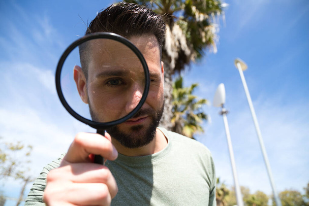 Handsome young man looks through a magnifying glass. His magnified eye can be seen from under the magnifying glass. The photo is taken from below and the man above. Concept detectives, view. - Foto, Bild