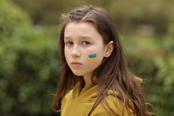 the face of a frightened girl, painted on her cheek in the yellow-blue colors of the Ukrainian flag, a request for help. Children ask for peace. High quality photo. High quality photo - 写真・画像