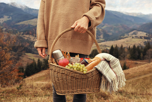Woman holding wicker picnic basket with thermos, snacks and plaid in mountains on autumn day, closeup - Fotografie, Obrázek
