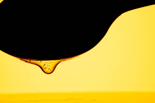 Thick drops of honey flowing down from a dark black spoon on yellow background. Golden streams of honey molasses or syrup are spilled. Sweet honeyed nectar dripping from a spoon close up. A product of - Photo, Image