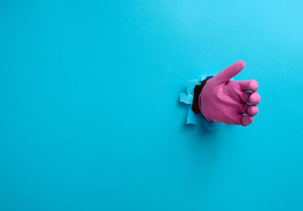 ruka v růžové latexové rukavici drží předmět, část těla vyčnívá z roztržené díry v pozadí z modrého papíru - Fotografie, Obrázek