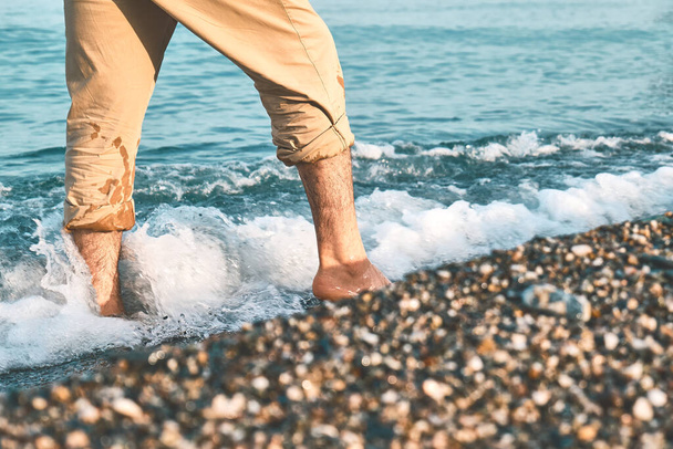 Man wearing shorts, walking barefoot along the seashore. Male legs walks on pebble beach along the shore near the water with waves, low section. Wellness, freedom and travel in summertime concept. - Photo, Image