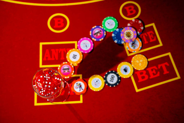 Top view of casino chips and a glass filled with red dice. A set of dice and playing chips for craps, poker, roulette on a red gaming table in a casino. Close up. Background of gambling, entertainment - Foto, Imagem