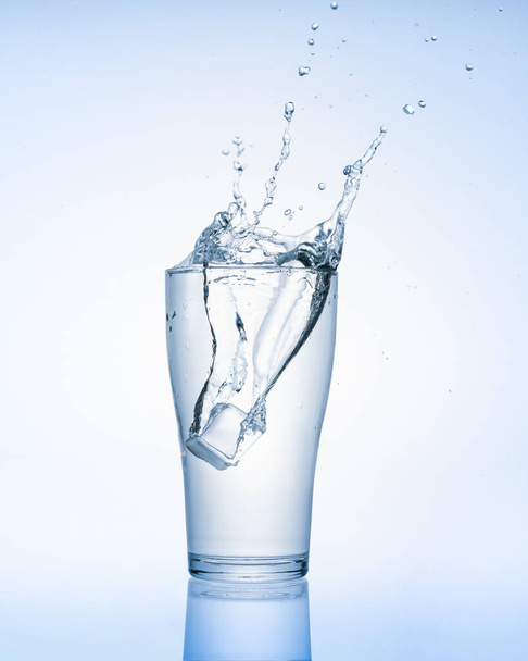 Cubo de hielo que cae con salpicaduras en agua de vidrio aislado sobre fondo blanco. Bebida saludable y estilo de vida. - Foto, imagen
