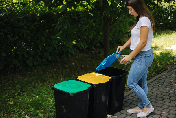 Portrait woman throwig empty glass bottle in recycling bin. Different colour of plastic bins in the park. - Foto, afbeelding