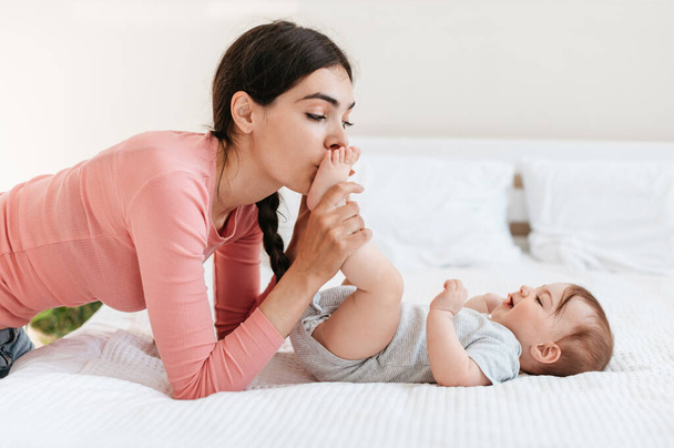 Οι μητέρες νοιάζονται. Αγαπημένη μητέρα να φιλάει μικροσκοπικά πόδια από το μωρό της, να απολαμβάνει το δέσιμο με το μωρό στο σπίτι, ξαπλωμένη στο κρεβάτι και να περνάει χρόνο μαζί - Φωτογραφία, εικόνα