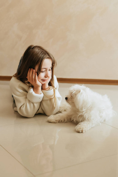 fille 9 ans avec des cheveux longs modèle avec un chien de compagnie écolière maltaise à la maison style de vie sur un fond beige allergie vétérinaire - Photo, image