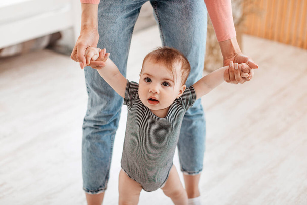 Little baby boy or girl taking first steps with mother help, woman holding childs hands, home interior, closeup. Cute kid daughter or son walking by bedroom - Zdjęcie, obraz