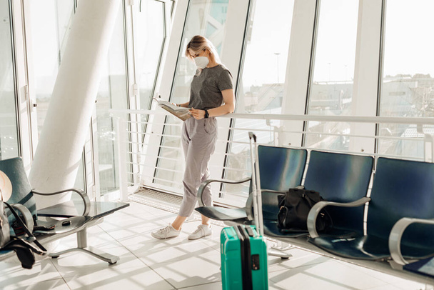 Portrait of woman wearing grey T-shirt, trousers, white disposable mask, standing near turquoise suitcase, reading newspaper in transit lounge of airport, waiting for flight. Travelling, safety. - Foto, imagen