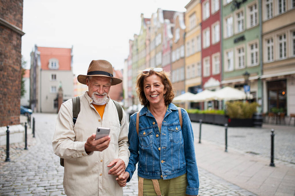A portrait of happy senior couple tourists smiling, holding hands, using smartphone outdoors in historic town - Foto, imagen