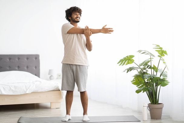 Cheerful millennial indian man in homewear practicing yoga at home, standing on fitness mat and stretching arms, looking at copy space and smiling, enjoying morning workout, panorama - Foto, imagen