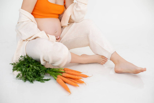 Обрізана фотографія вагітної жінки, що сидить з пучком апельсинових овочів, зеленою морквою. Носіть лляний одяг. Тримає ретельно живіт. Свіже харчування дієта, здорове харчування. Білий фон
 - Фото, зображення