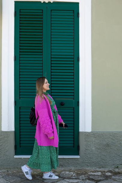 Vertical image of woman looking away in elegant dress and jacket, staying against big louver green door entrance. Walking like traveler while visiting foreign country. Side view of radial path - Zdjęcie, obraz